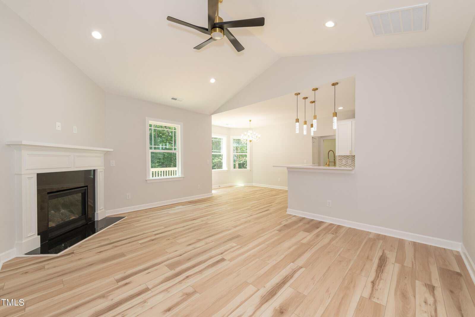 Spacious, modern living area with light wood floors, a ceiling fan, a fireplace, and an open layout leading to a bright kitchen with pendant lights and large windows.