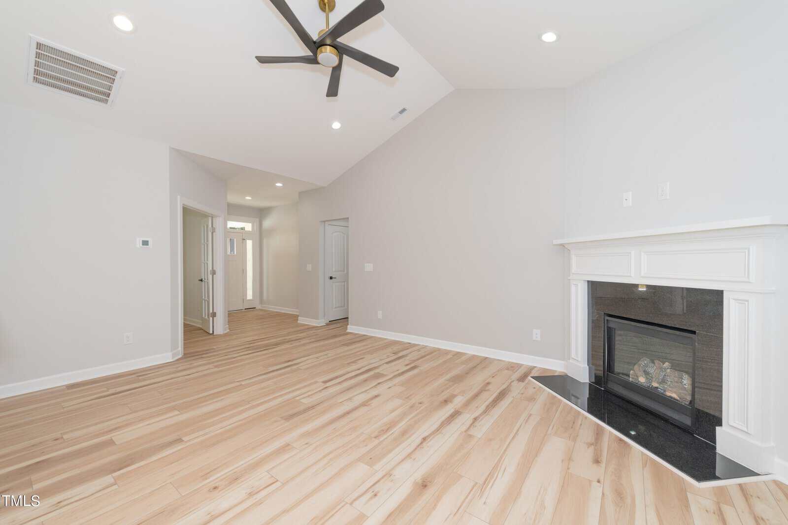 A spacious, empty living room with light wood flooring, a white fireplace with black trim, light gray walls, recessed lighting, and a black ceiling fan. A hallway and doors are visible in the background.