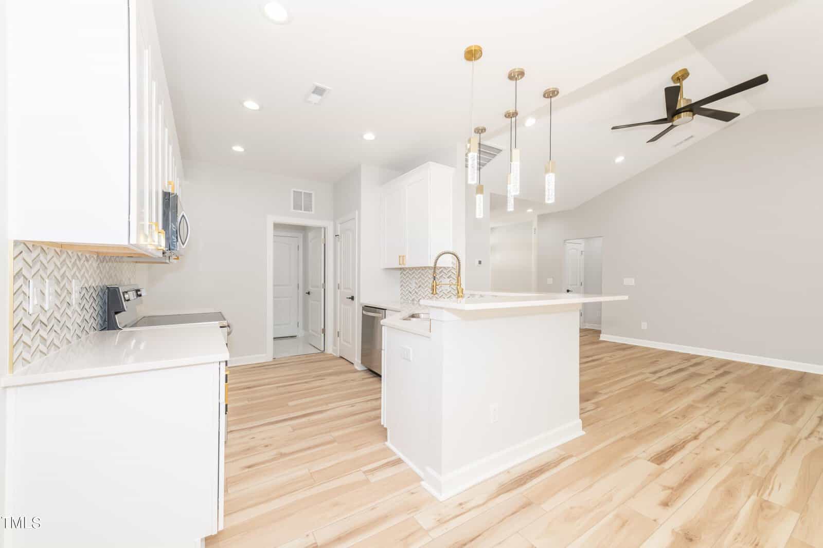 A modern kitchen with white cabinets, stainless steel appliances, light wood flooring, a white island with pendant lights above, and a ceiling fan in an open, bright space.