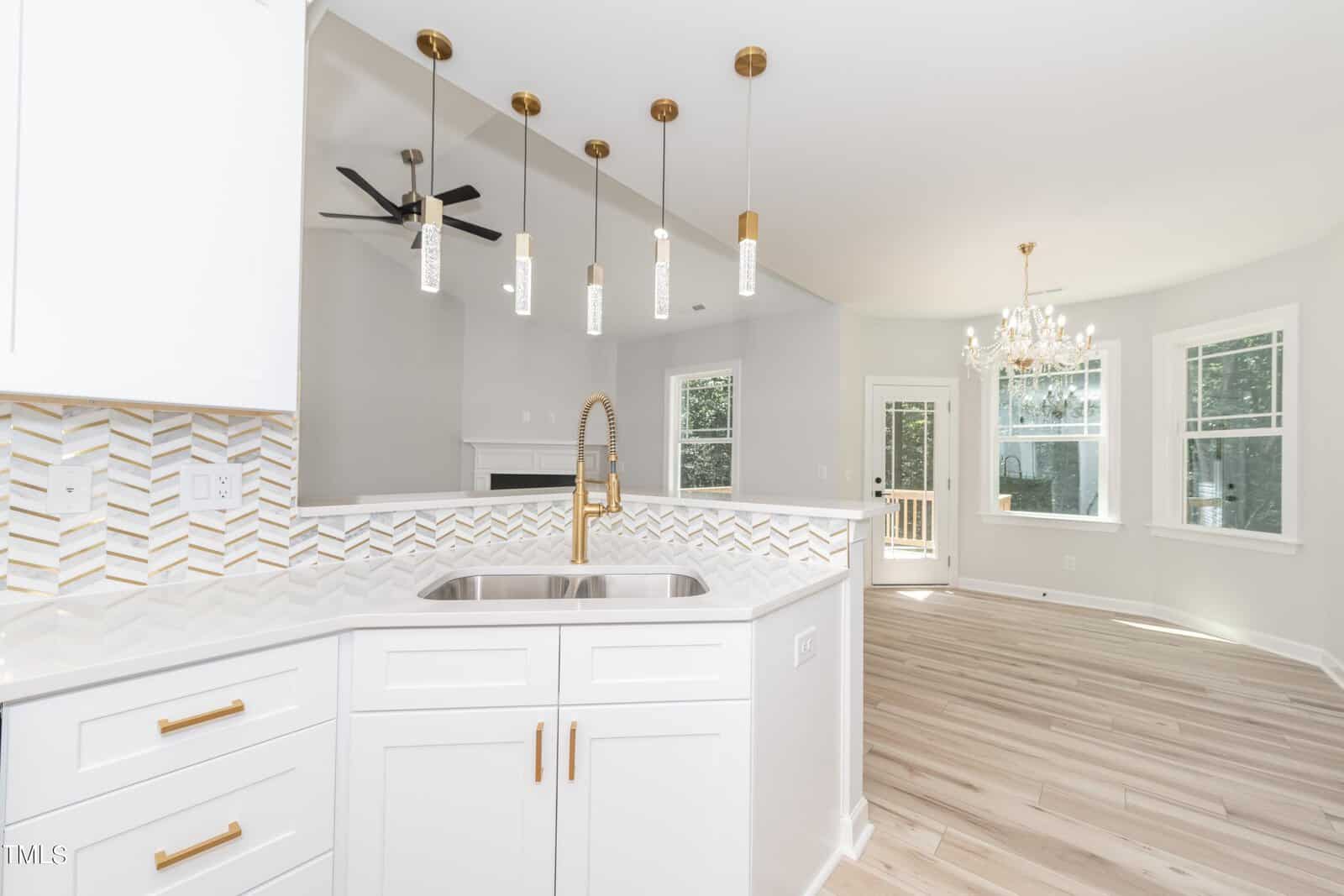 Modern kitchen with white cabinets, gold handles, and geometric backsplash. Pendant lights hang above a sink on a peninsula counter. Open layout leads to a bright living and dining area with large windows and a chandelier.