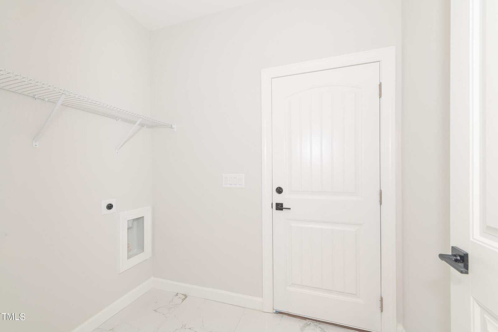 A laundry room with white walls, a tile floor, a white door, a wire shelf, and hookups for a washer and dryer. The room is empty and brightly lit.