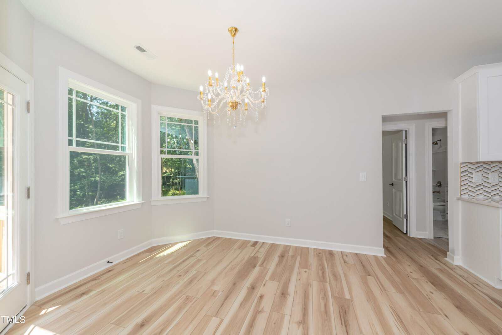Bright, empty room with light wood floors, white walls, two windows, a glass door, and a gold chandelier. An open doorway reveals another room and a glimpse of a bathroom. Natural light fills the space.