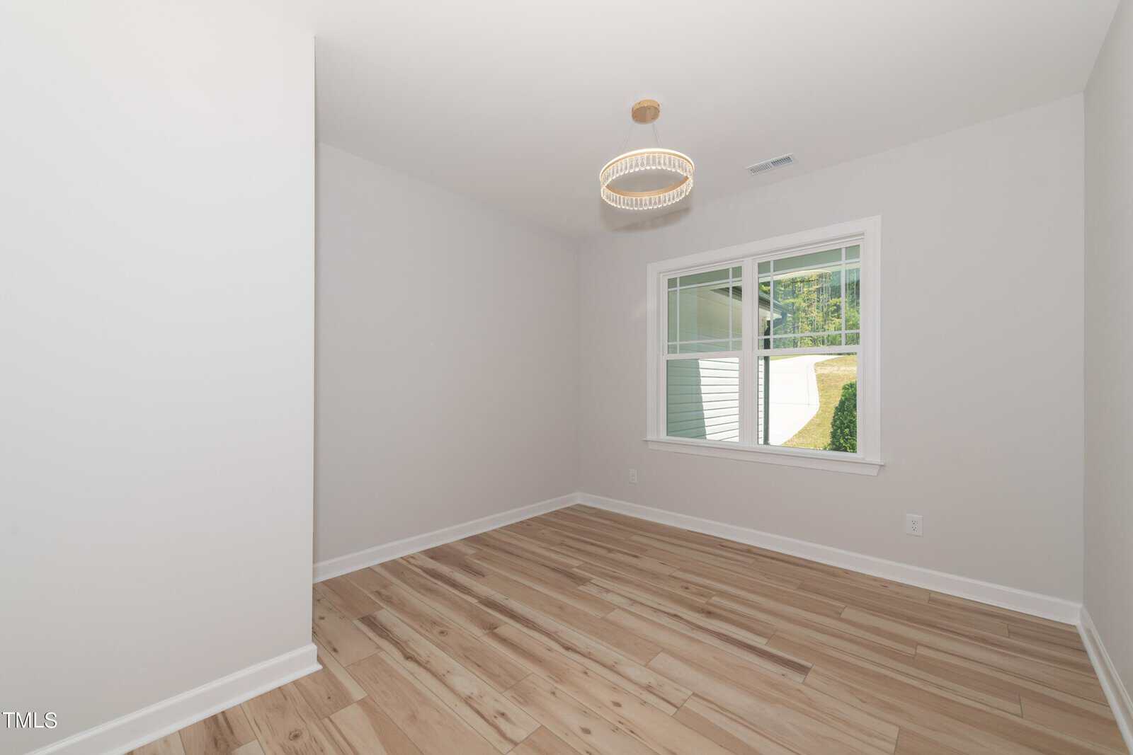 A small, empty room with light-colored wood floors, white walls, a window with white trim, and a modern round light fixture on the ceiling. Natural light enters through the window.