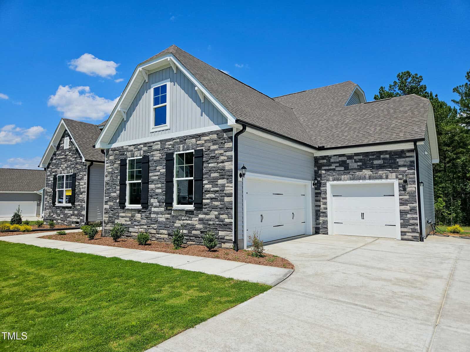 A modern house with gray stone and white siding exterior, two white garage doors, manicured lawn, small shrubs, and a clear blue sky overhead.
