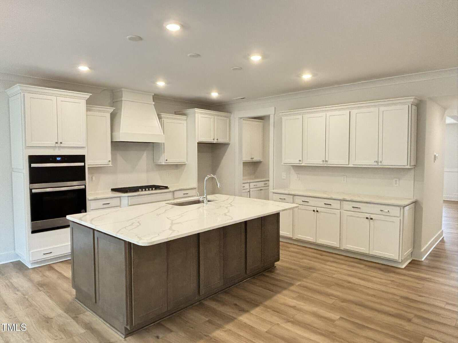 Modern kitchen with white cabinets, wood flooring, a large island with a marble countertop, built-in oven, stovetop, and overhead lights. The kitchen is clean, spacious, and unfurnished.
