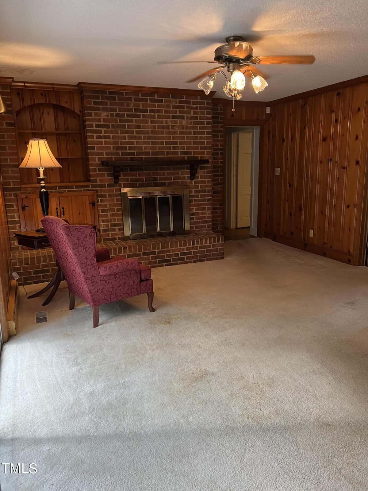 A cozy living room with a brick fireplace, wooden paneling on the walls, and beige carpet. A maroon armchair is placed next to a wooden side table with a lamp. A ceiling fan with lights hangs from the white ceiling. Shelving is built into the wall near the fireplace.