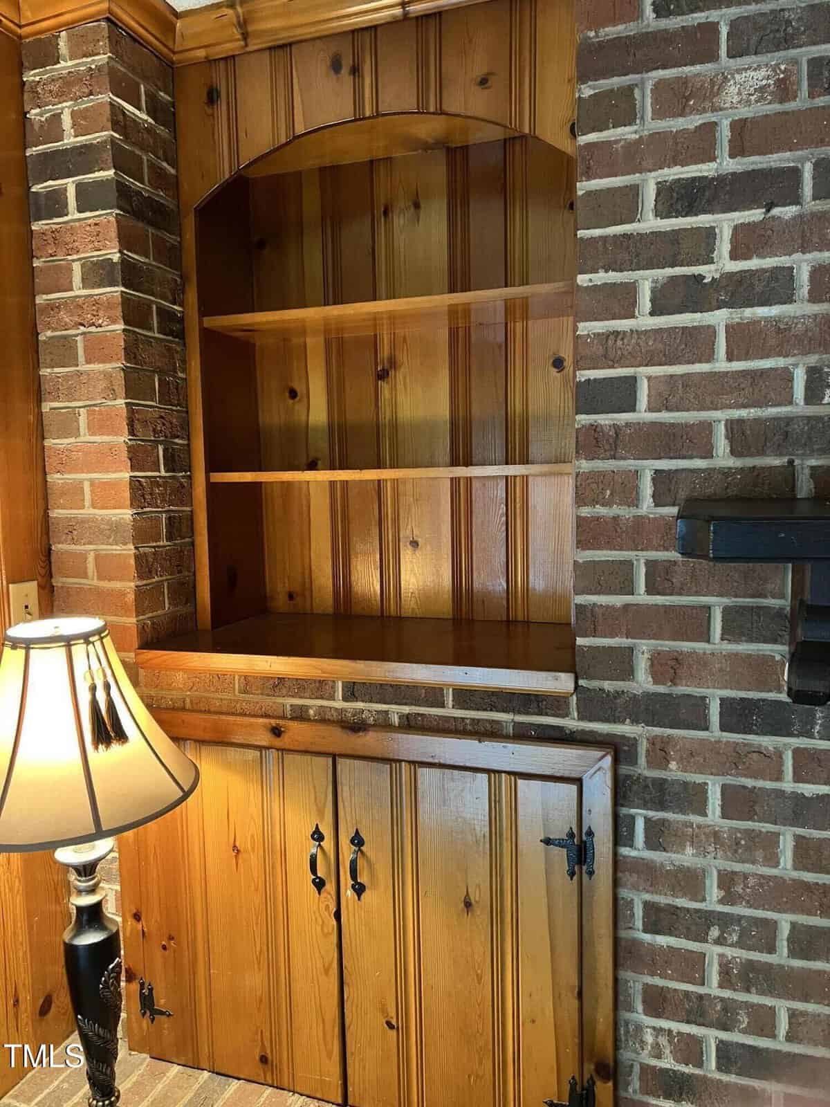 A wooden built-in shelving unit is set within a brick wall. The lower section features closed cabinet doors, while the upper section has open shelves. A lamp with a beige shade stands next to the unit. The setting appears to be a cozy indoor space.