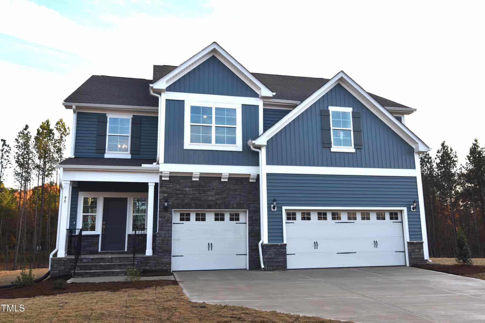 A two-story blue house with white trim, three garage doors, a covered front porch, and a concrete driveway. The house has stone accents and is surrounded by trees and an unfinished lawn.