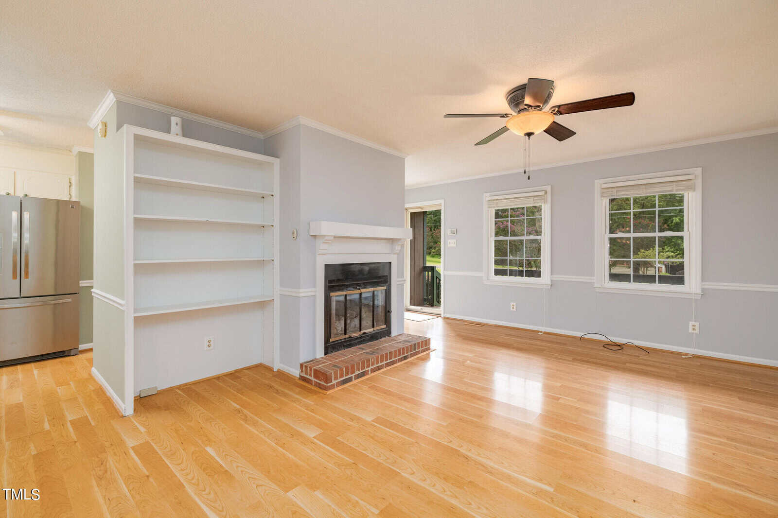 A bright living room with light wooden flooring features a built-in white bookcase, a brick fireplace with a mantel, and two large windows. There's a ceiling fan with a light fixture and an open doorway leading to a modern kitchen with stainless steel appliances.