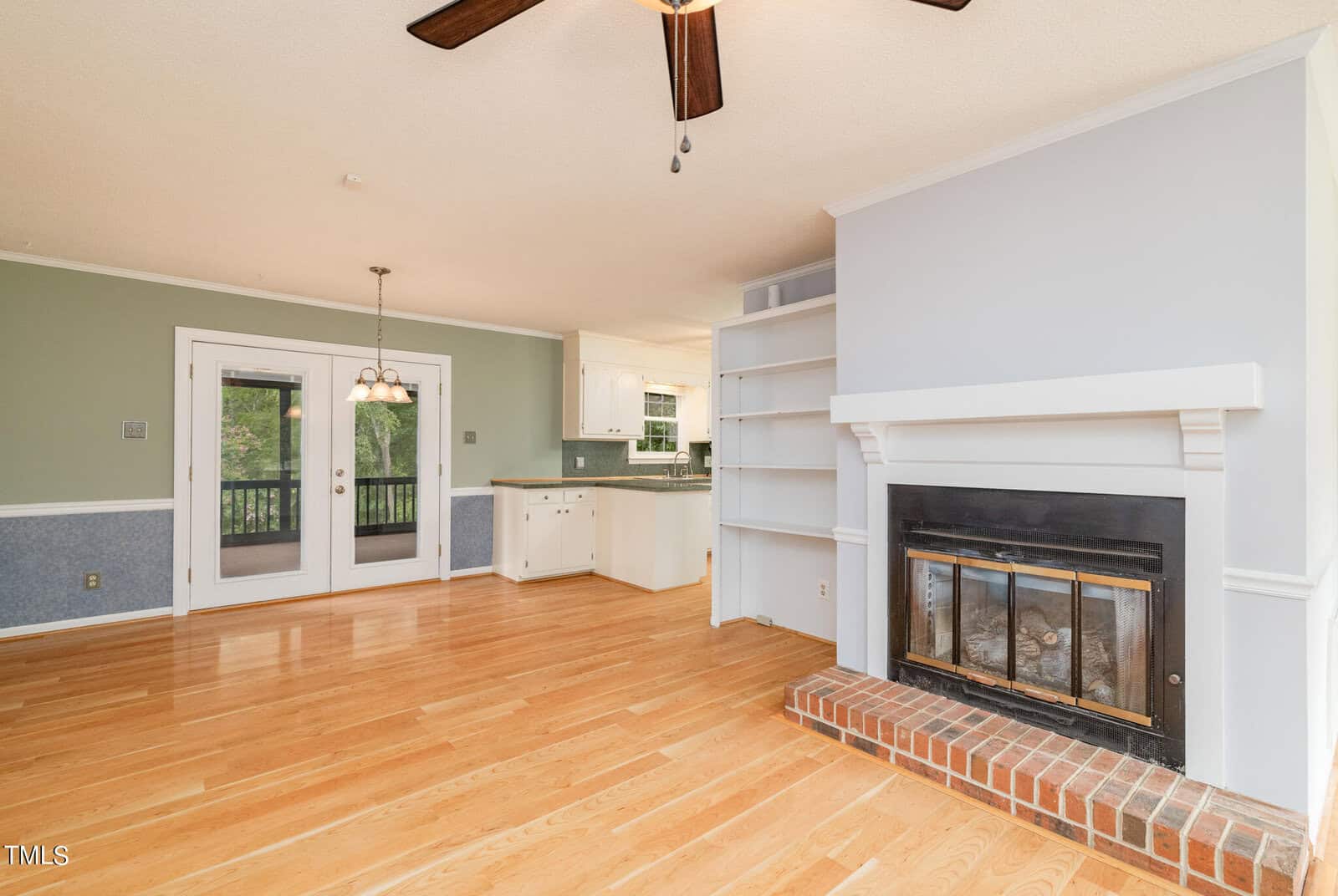 A spacious living area with polished hardwood floors, a built-in fireplace with a brick base, white shelving units, and a ceiling fan. In the background, there's a dining area next to a double-door exit to the outside, and a view of a kitchen with white cabinets.