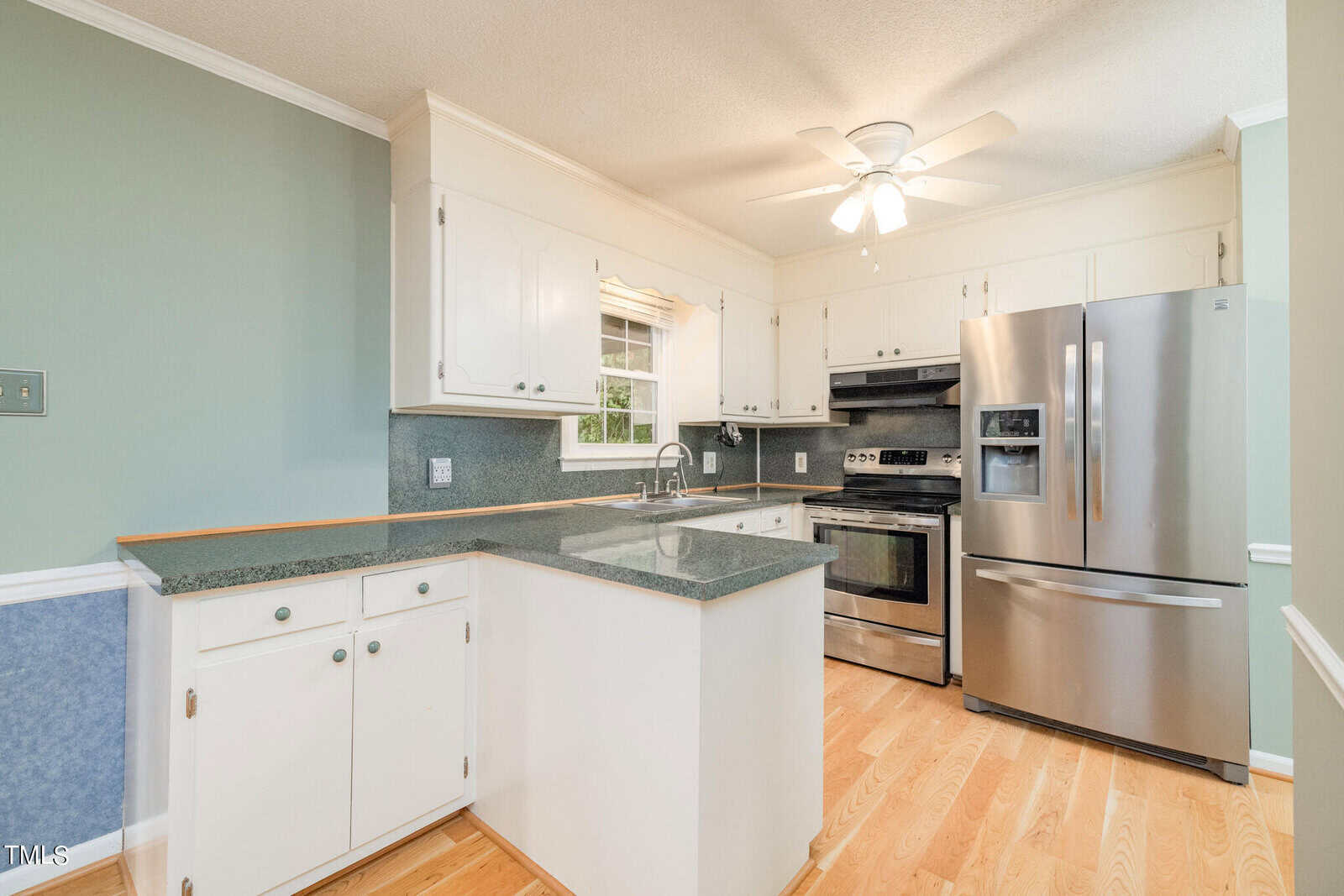 A kitchen with white cabinetry, stainless steel appliances including a refrigerator, oven, microwave, and dishwasher. There is a double sink, green countertops, wooden flooring, sage green walls, and a ceiling fan with a light fixture above.