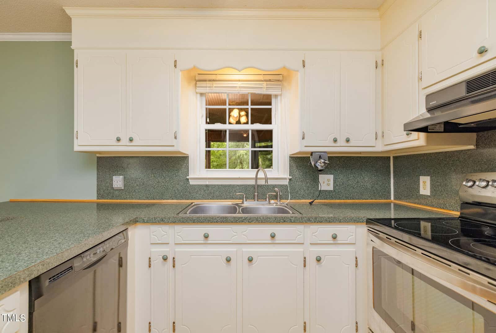 A kitchen with white cabinetry, a double sink under a window, green speckled countertops, and stainless steel appliances including a dishwasher and an oven. The window overlooks a green yard, and lights are reflected in the window's glass.