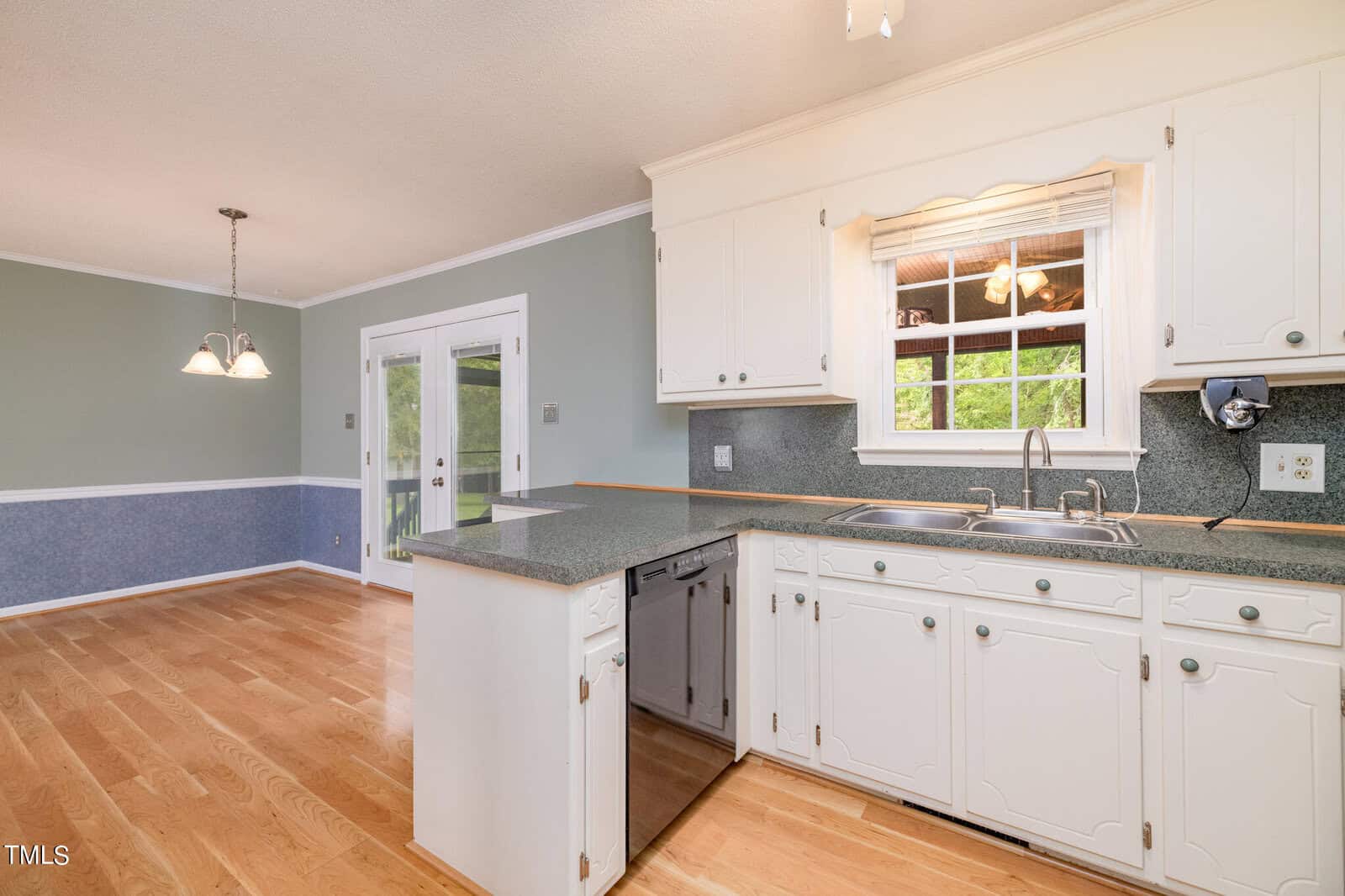 A kitchen with white cabinetry and green countertops opens into a dining area with light wood flooring and green walls. There is a double sink under a window and a dishwasher to the left. A pendant light fixture hangs above the dining space near glass patio doors.
