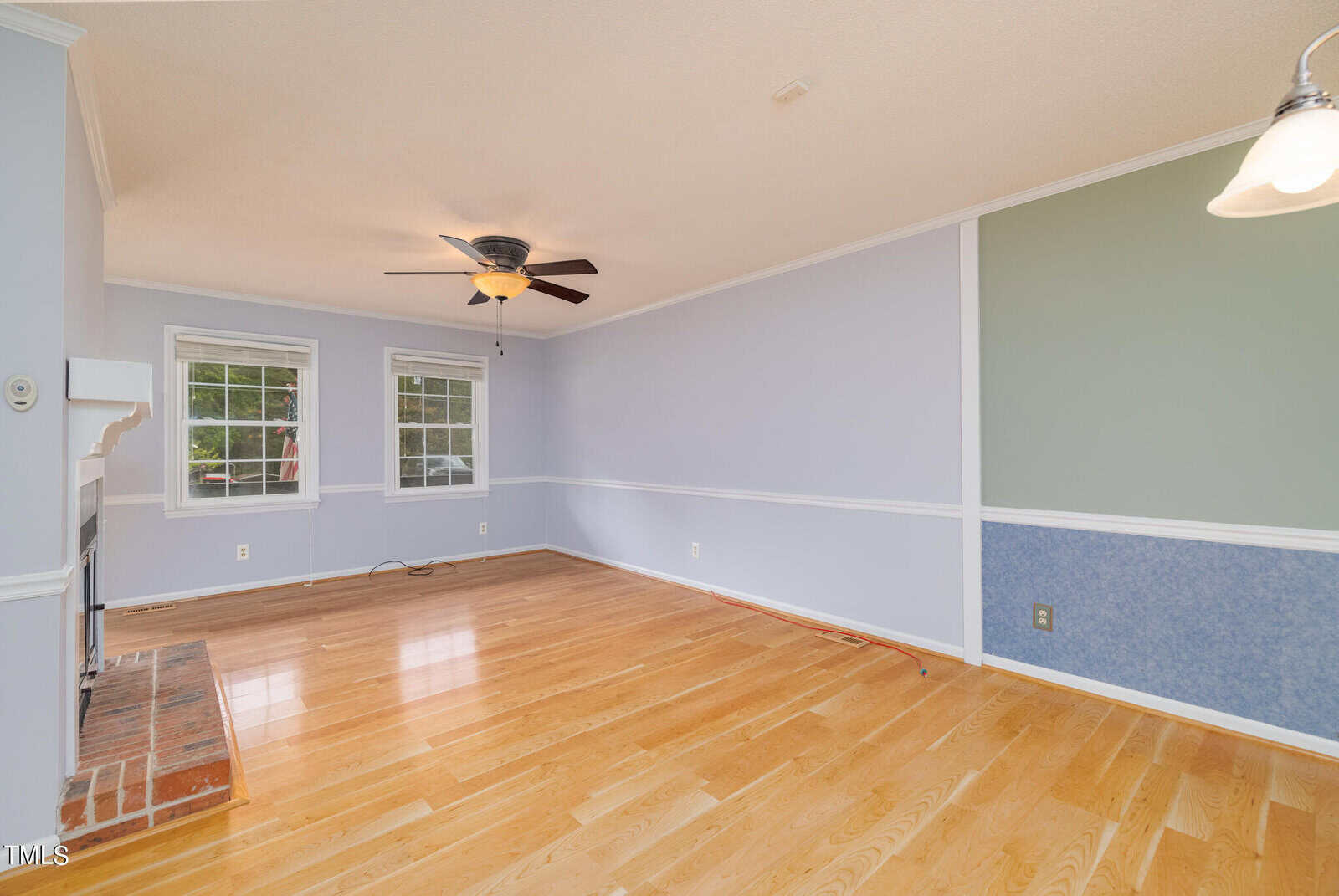 A spacious, empty living room with light wood flooring, light blue walls, crown molding, and two large windows. The room features a ceiling fan with a light, a partial brick fireplace on the left, and a mix of green and blue accent walls.
