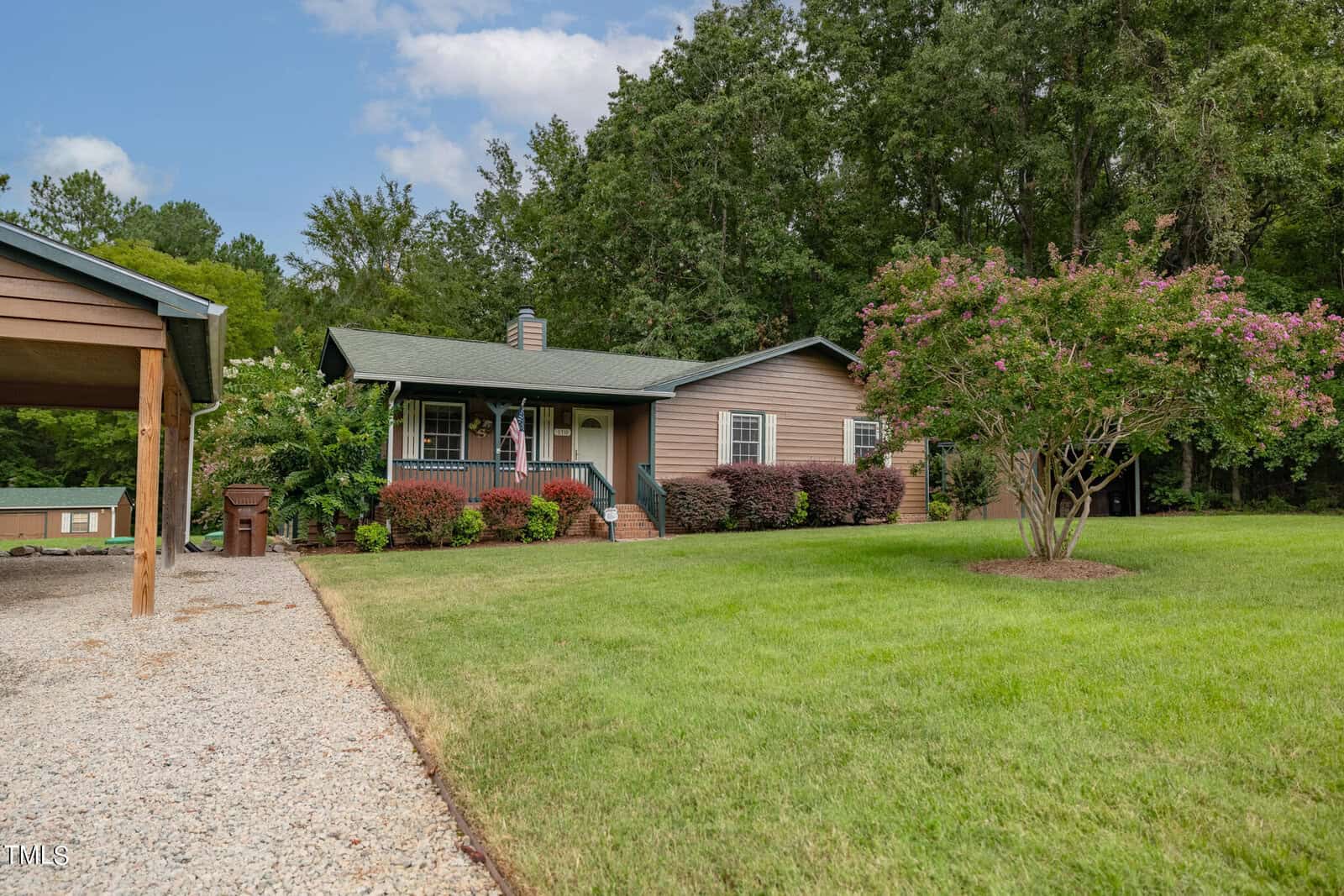 A charming single-story home at 110 Lake Road in Henderson features a green roof, beige siding, and a covered front porch. The house boasts a well-manicured lawn, gravel driveway, carport, and is surrounded by trees and colorful shrubs. A flowering tree graces the front yard under a partly cloudy sky.