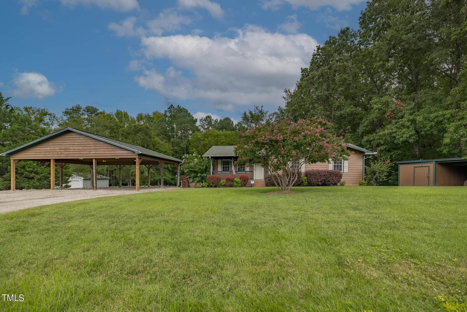 A single-story brown house with a green roof is surrounded by a lush green lawn and trees. To the left is a covered carport with space for several vehicles, and on the right is a shed. The sky above is partly cloudy.