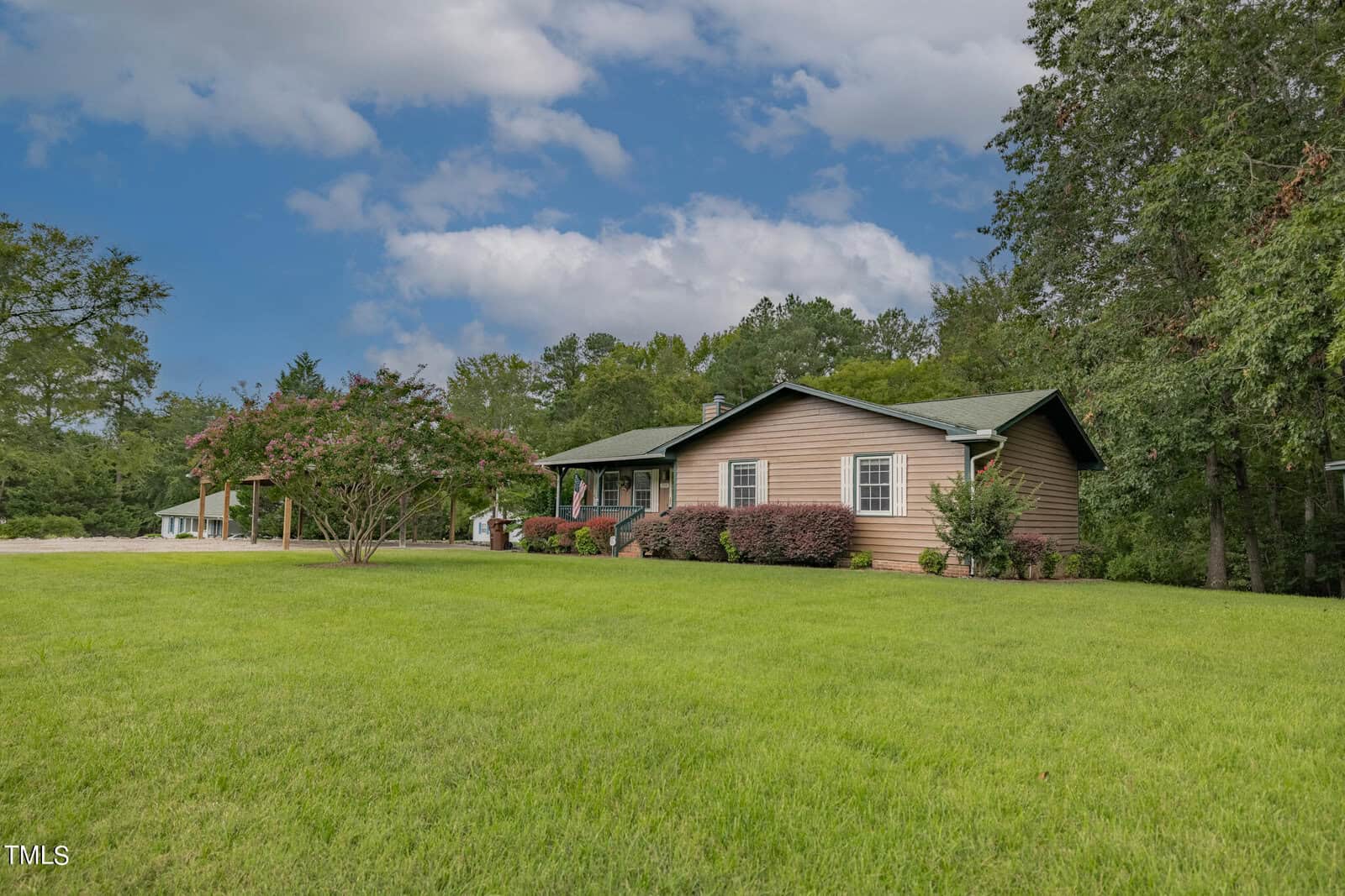A single-story, light brown house with a green roof, surrounded by well-maintained greenery and trees. The house has a covered front porch with chair and shrubs lining the walkway. The sky above is partly cloudy, and the expansive lawn is lush and green.