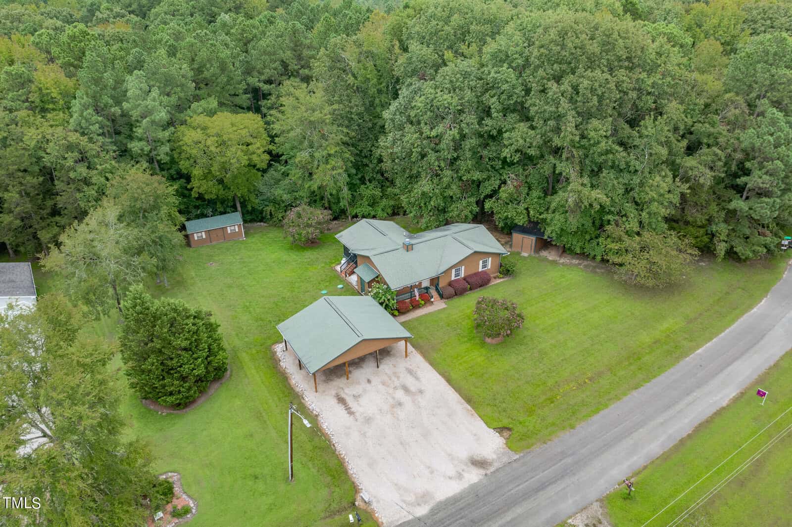 Aerial view of a rural residence surrounded by trees. The property features a house with a green roof and two outbuildings, a detached carport, and a gravel driveway with parking space. The house has a well-maintained front yard and a backyard adjoining a wooded area.
