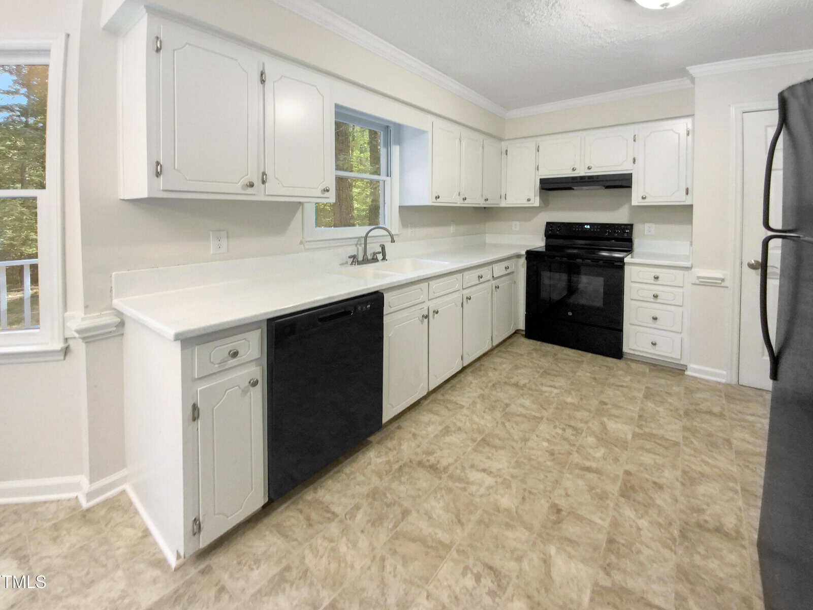 Bright kitchen with white cabinets, white countertops, a black stove, black dishwasher, and a large window above the sink. The floor has a light-colored patterned tile, and walls are painted off-white.