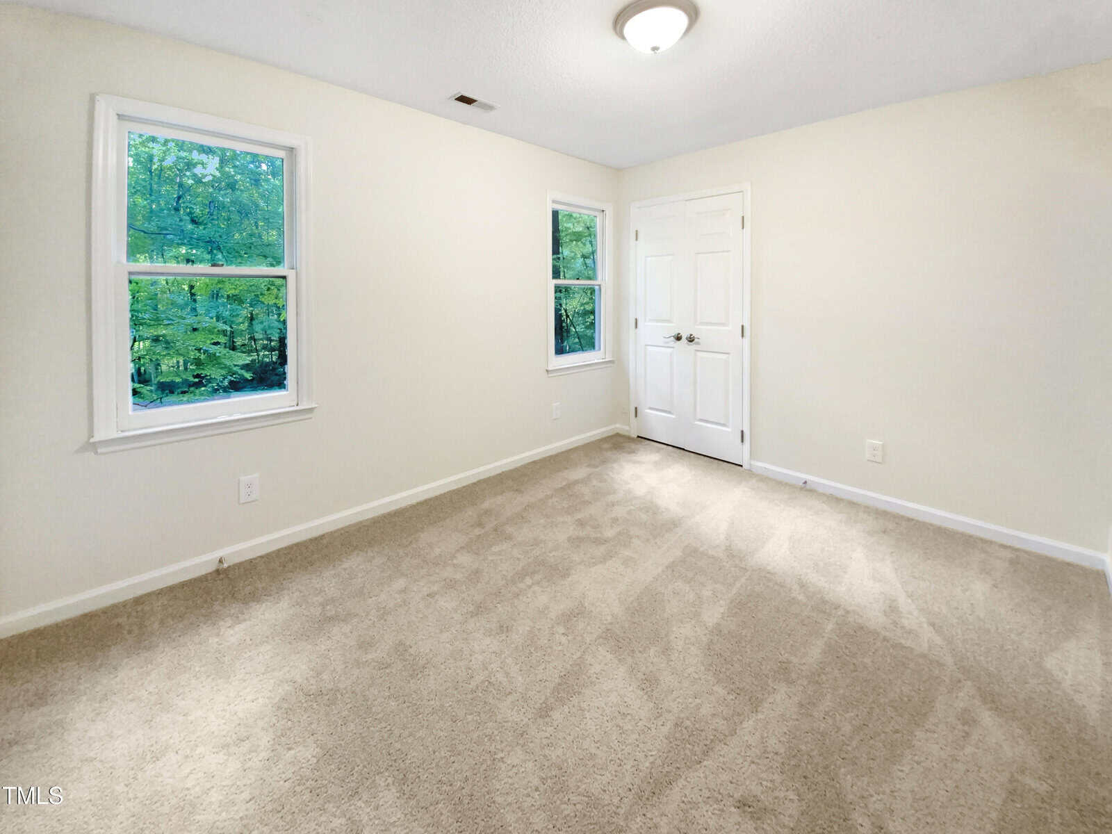 Empty room with beige carpet, white walls, two windows showing green trees outside, and a closed double door; ceiling light fixture is on, creating a bright, clean appearance.