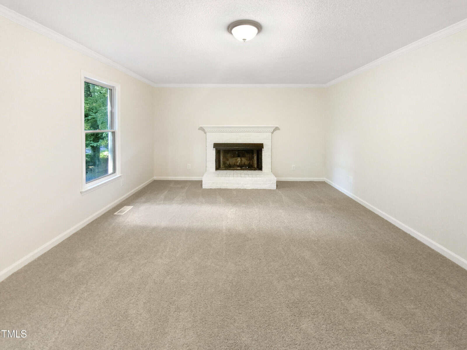 A bright, empty living room with beige carpet, cream-colored walls, a white ceiling, a central light fixture, a window on the left, and a white fireplace on the far wall.