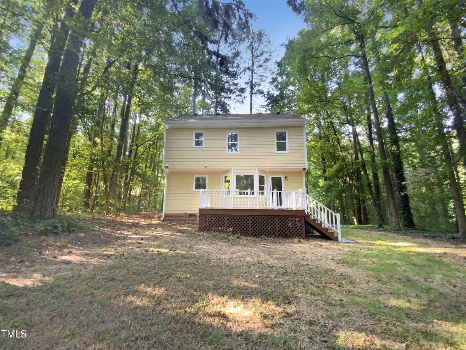 A two-story yellow house with white trim and a deck, surrounded by tall trees and greenery, sits on a grassy, shaded lot.