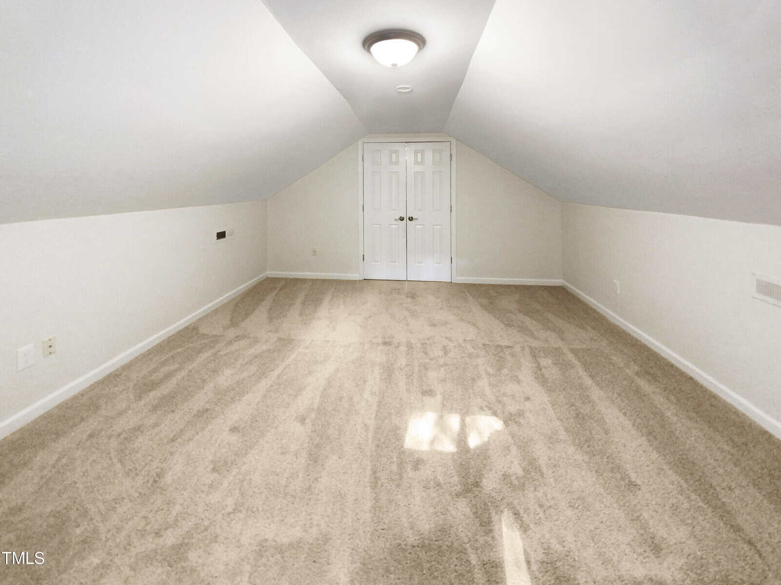 A spacious, empty attic room with sloped ceilings, beige carpet, white walls, and double white doors at the far end under a ceiling light. Sunlight shines through a small window out of view.