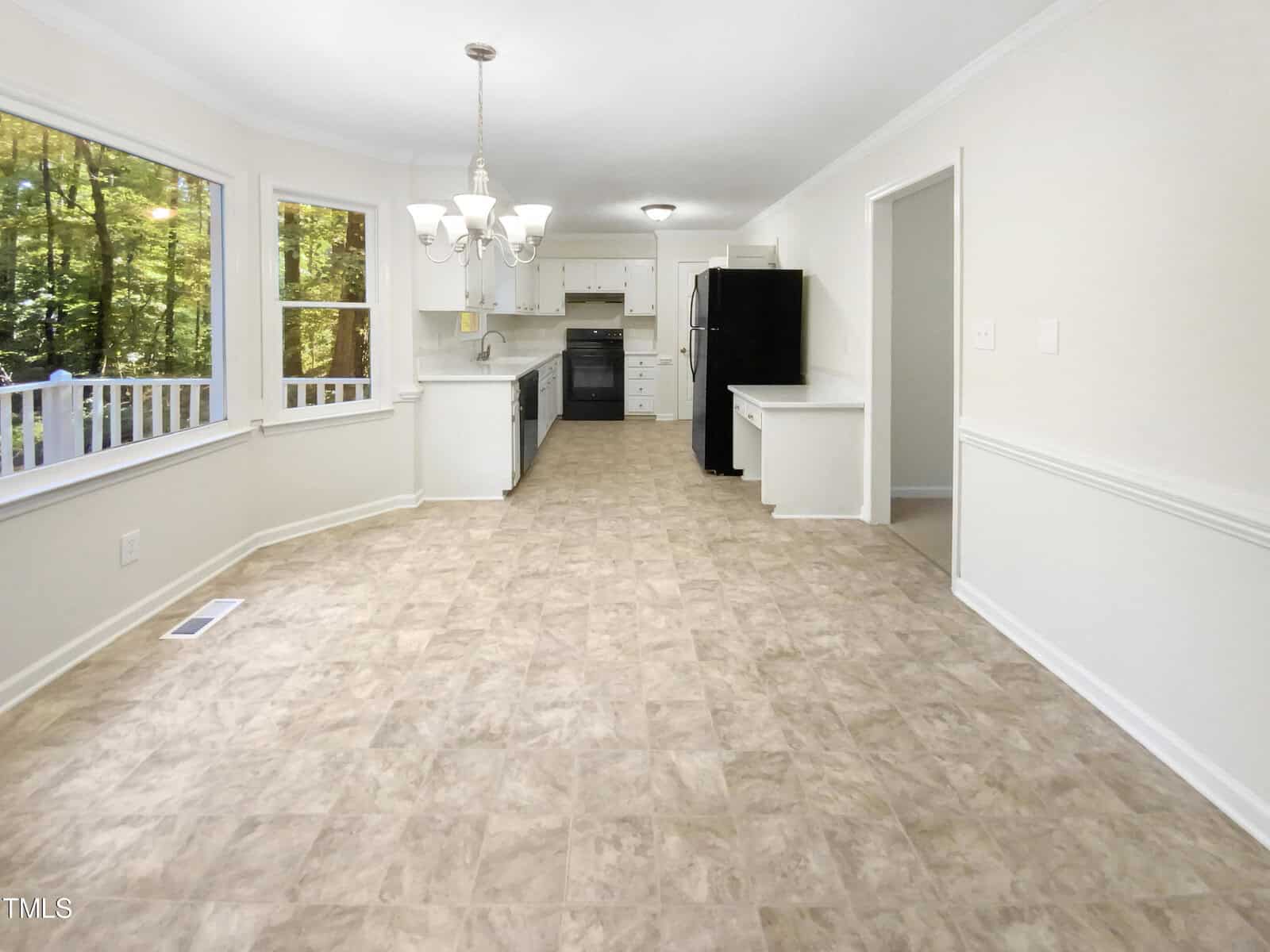 Bright, open kitchen and dining area with large windows, white cabinets, black appliances, a chandelier, and beige tile flooring. A deck and green trees are visible outside the windows.