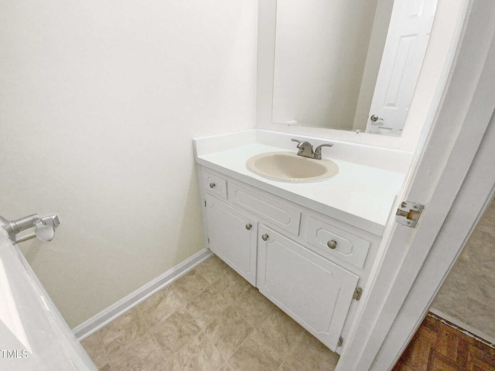 A small bathroom with a white vanity, beige sink and faucet, large mirror, white cabinets, and light-colored tile flooring. The door is slightly open, showing part of a wooden floor outside.