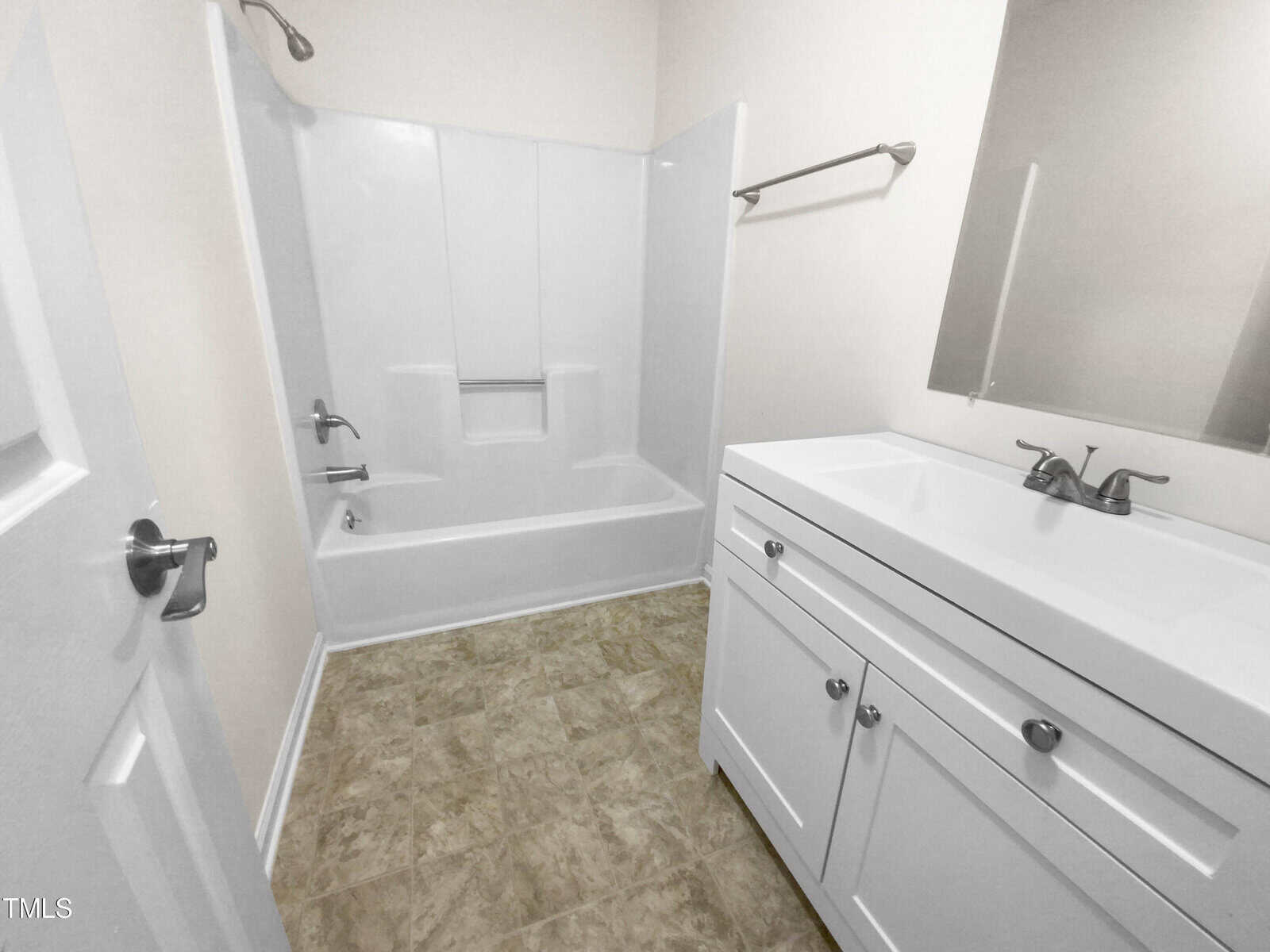 A clean, modern bathroom with a white bathtub and shower combo, a white vanity with dual faucets, a large mirror, towel racks, and beige tile flooring.