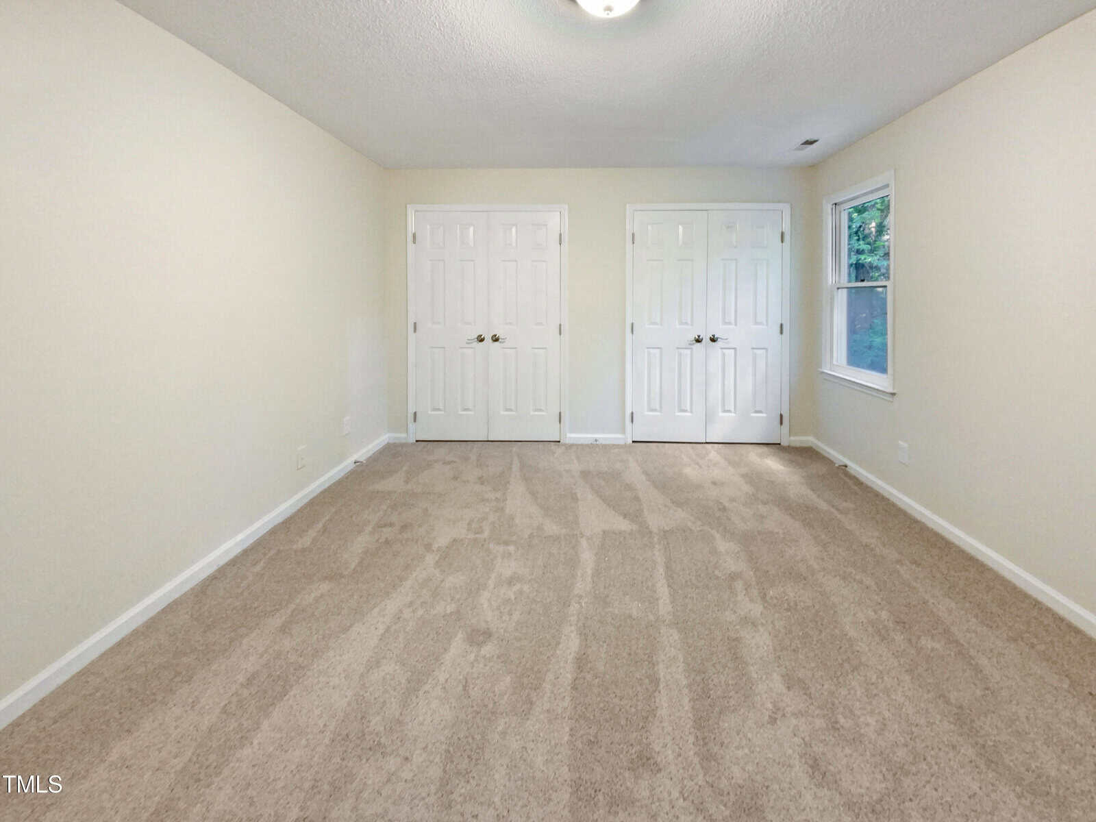 A bright, empty room with beige carpet, cream-colored walls, two double-door closets side by side, and a window letting in natural light.