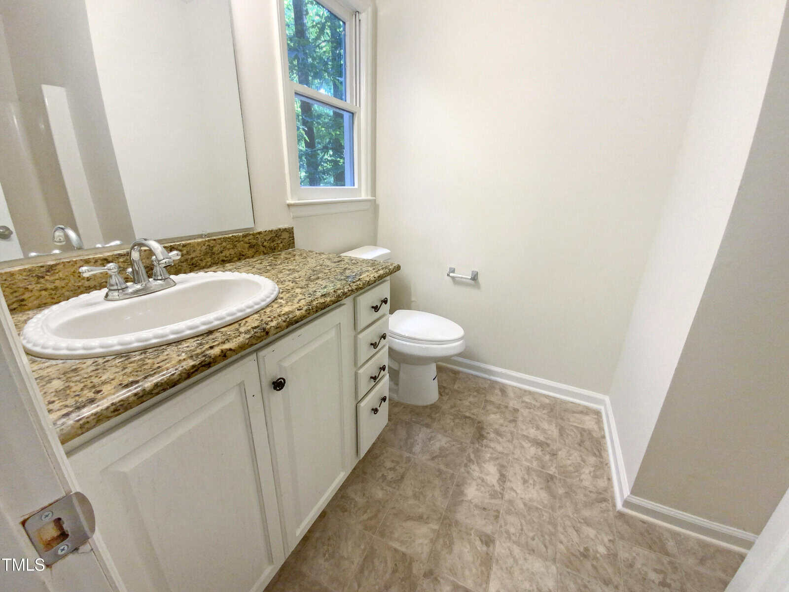 A bathroom with a granite countertop, white sink, white cabinets, a toilet, wall-mounted toilet paper holder, and a window letting in natural light.