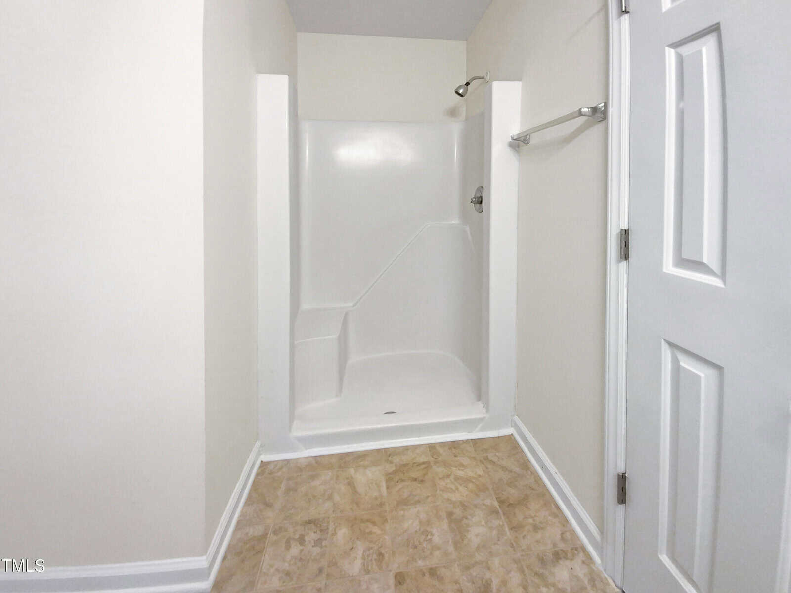A white walk-in shower with built-in seat, towel bar on the right wall, beige tile floor, and a white door partially visible.