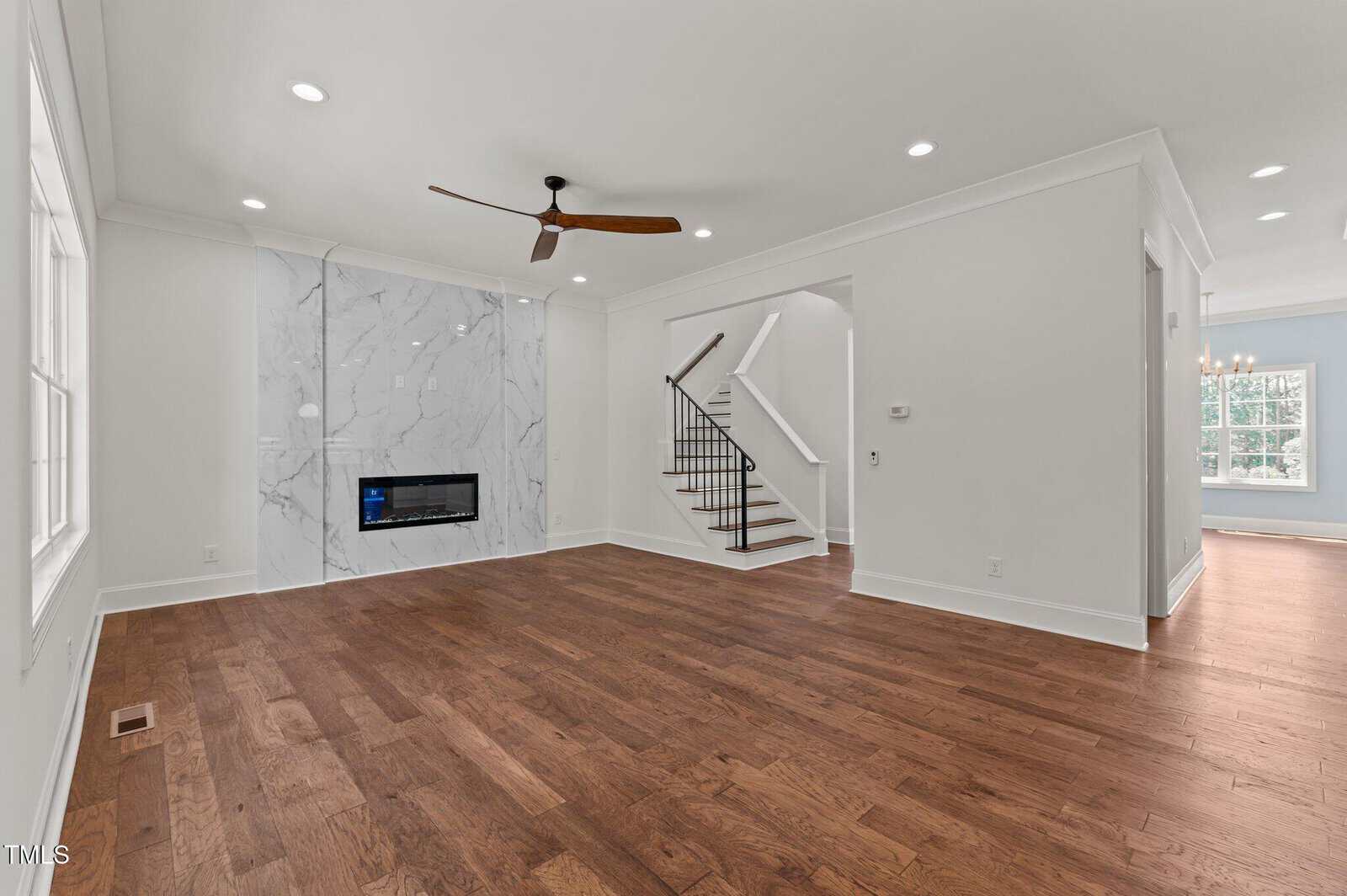 Spacious living room with wood floors, a marble accent wall with a modern fireplace, ceiling fan, white walls, and an open staircase leading upstairs; large windows bring in natural light.