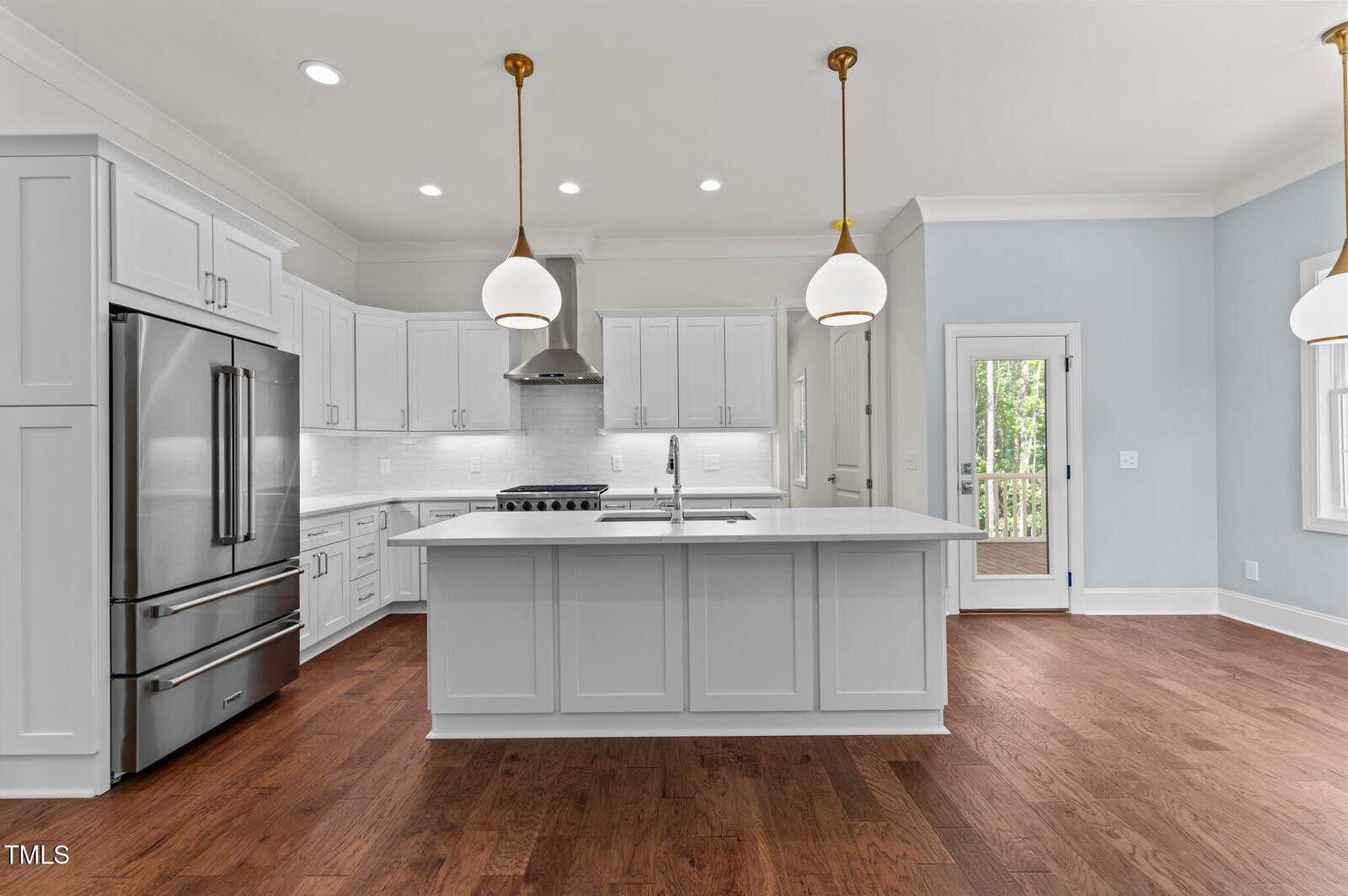 Modern kitchen with white cabinets, stainless steel appliances, large center island with sink, pendant lights above, wood flooring, and a door leading outside; space is bright with natural and overhead lighting.