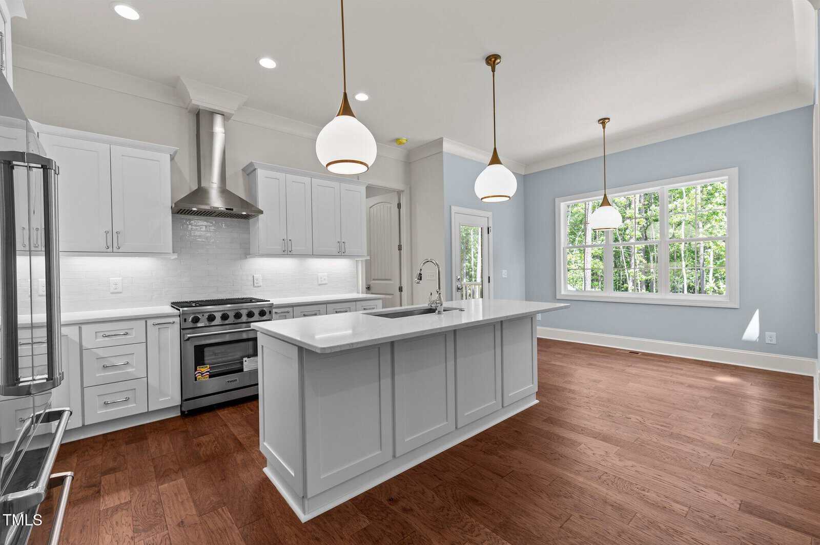 Modern kitchen with white cabinets, stainless steel appliances, large island with sink, pendant lights above, and an adjacent dining area with large windows and hardwood floors.