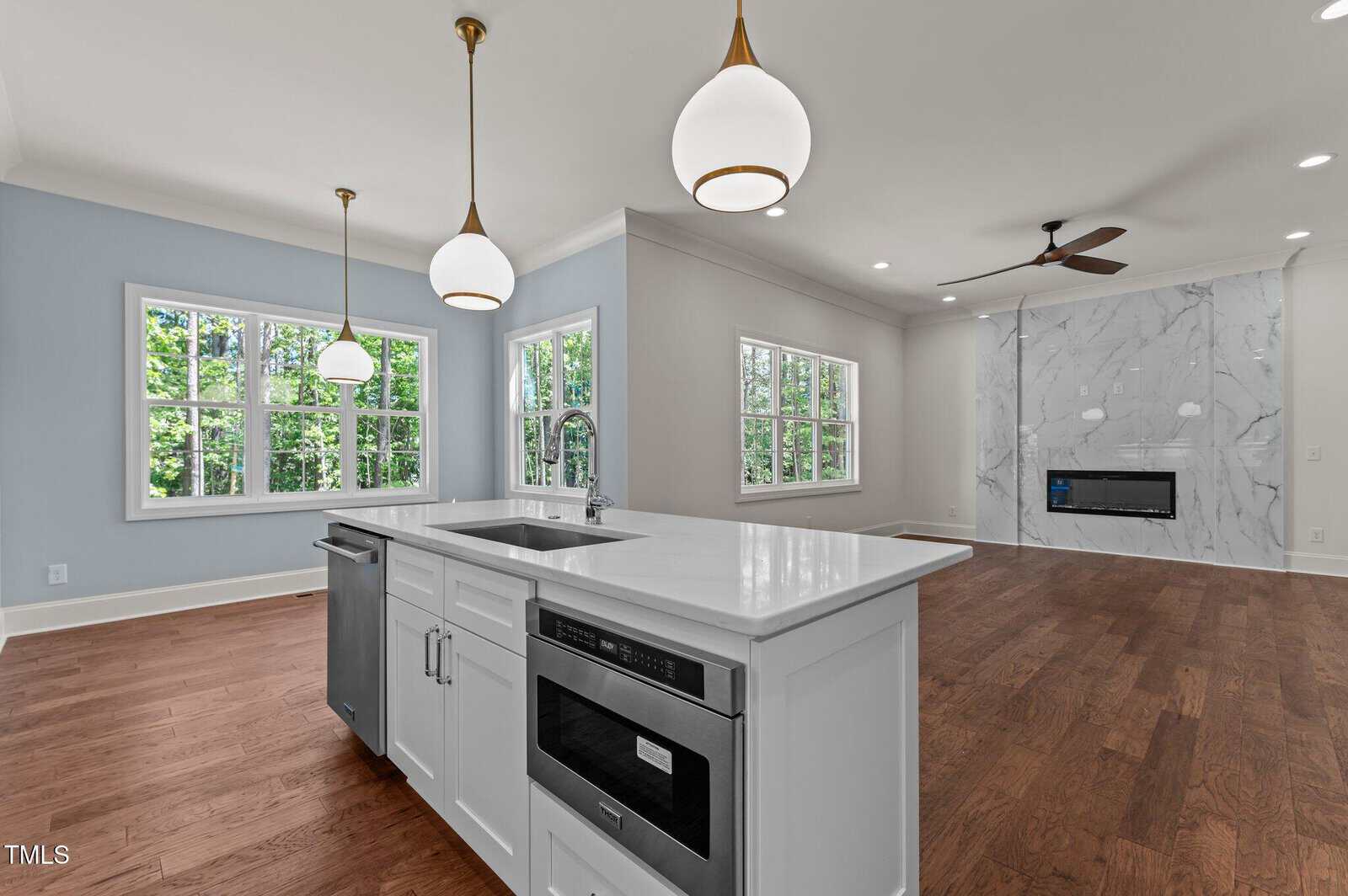 A modern, bright kitchen with a white island, built-in microwave, and pendant lights, open to a living area with wood floors, large windows, and a marble accent wall with a fireplace.