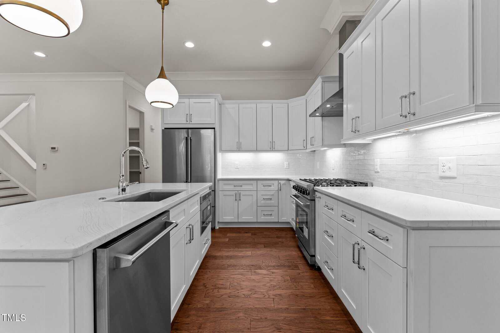 Modern white kitchen with stainless steel appliances, marble countertops, dark wood floors, pendant lights, and sleek cabinetry. The space is clean, bright, and open with under-cabinet lighting highlighting the backsplash.