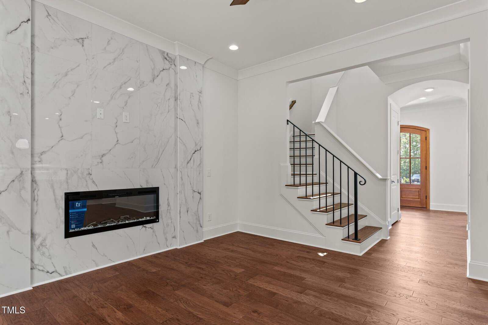 Living room with wood floors, a marble accent wall featuring a modern electric fireplace, white walls, recessed lighting, open staircase with black railing, and a wooden front door visible in the background.