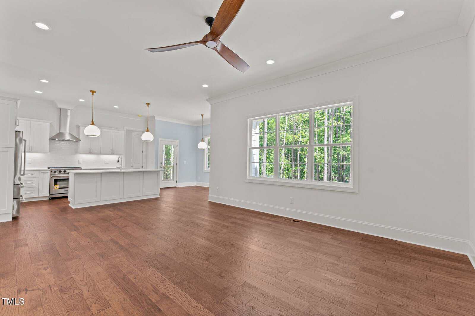 Spacious open-concept kitchen and living area with hardwood floors, large windows, white cabinetry, pendant lighting over a kitchen island, and a modern ceiling fan. Natural light fills the room.