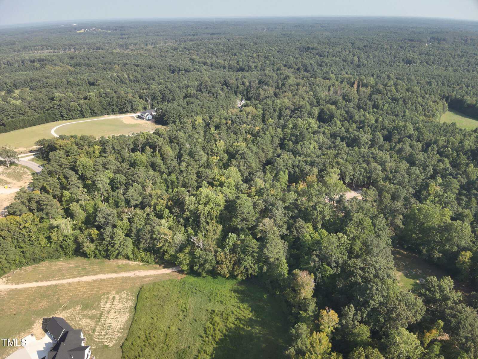 Aerial view of a densely wooded area with green trees, a dirt path, and a cleared section near the bottom left corner next to part of a house. A winding road and open grassy field are visible in the background.