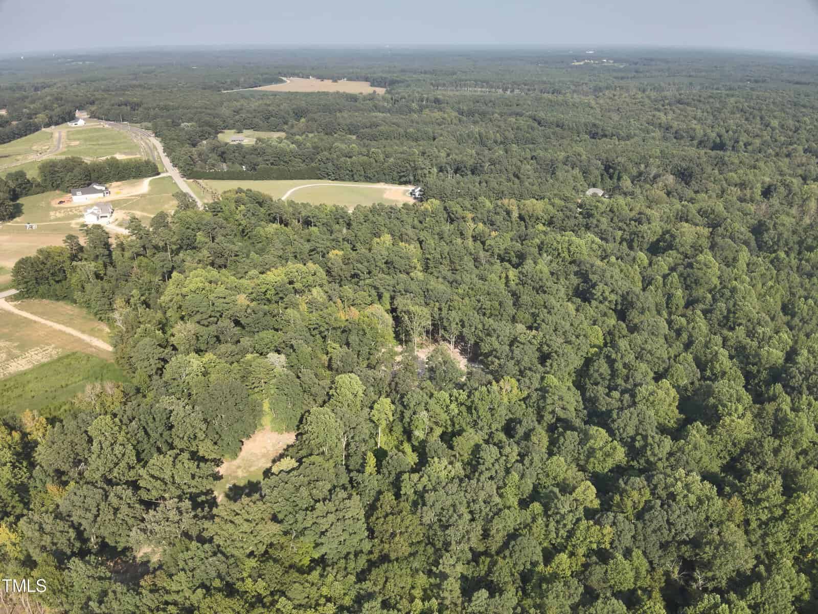 Aerial view of a densely wooded area with surrounding open fields, a few scattered houses, and roads cutting through the landscape on a clear day.
