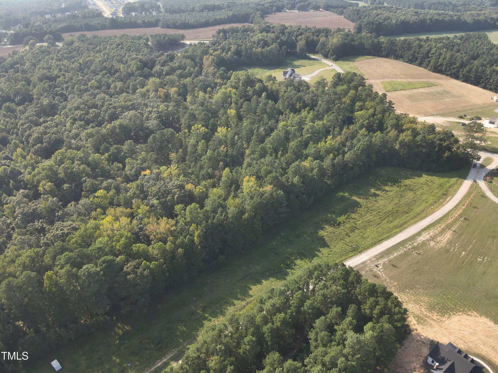 Aerial view of a wooded area bordered by open fields, a few houses, and winding roads. The landscape includes dense green trees, grassy clearings, and patches of tilled land.