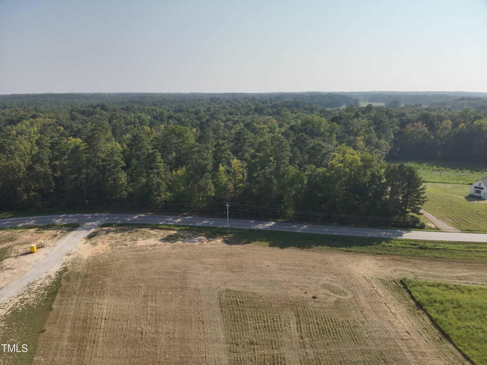 An aerial view of a cleared, grassy lot bordered by a two-lane road and dense forest. A single white house and open field are visible in the distance under a clear sky.