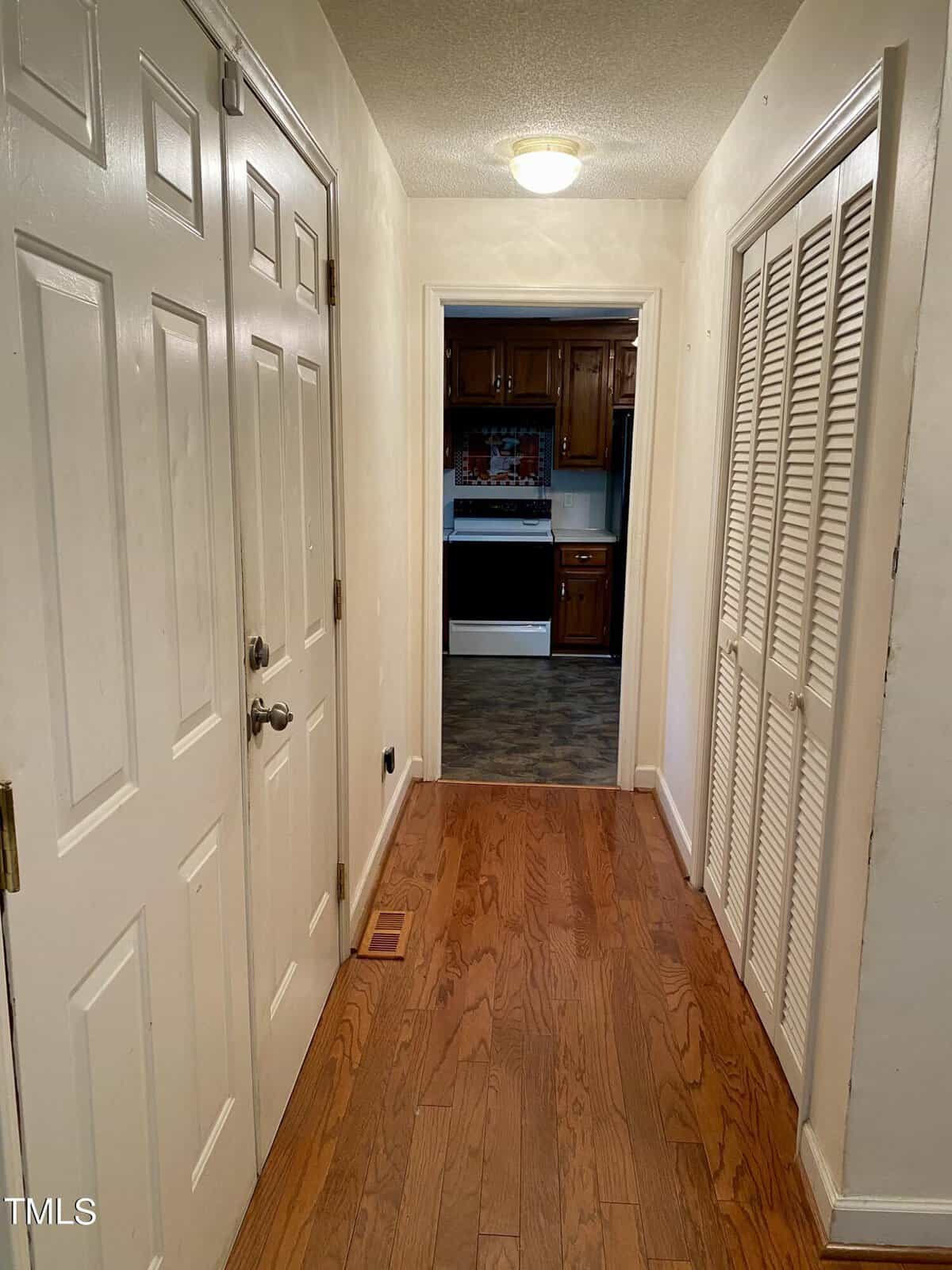 A hallway with light-colored walls and wooden flooring. On the left, there is an entry door with double locks. To the right, there's a set of white louvered closet doors. The hallway leads to a kitchen with dark wood cabinets and a stove visible.