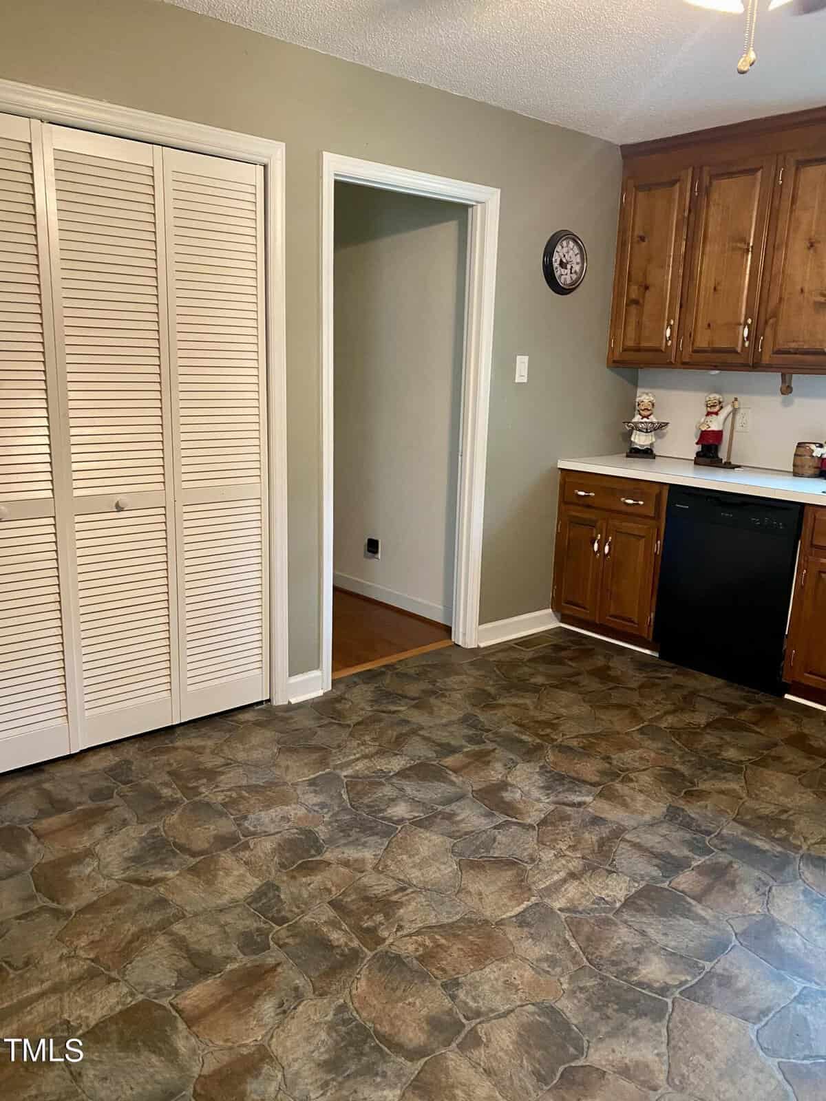 A kitchen with stone tile flooring, wooden cabinets, and white countertops. There is a doorway to an adjacent room, a double-door pantry on the left, and a dishwasher under the counter. A wall clock and small decorative items are on the counter.