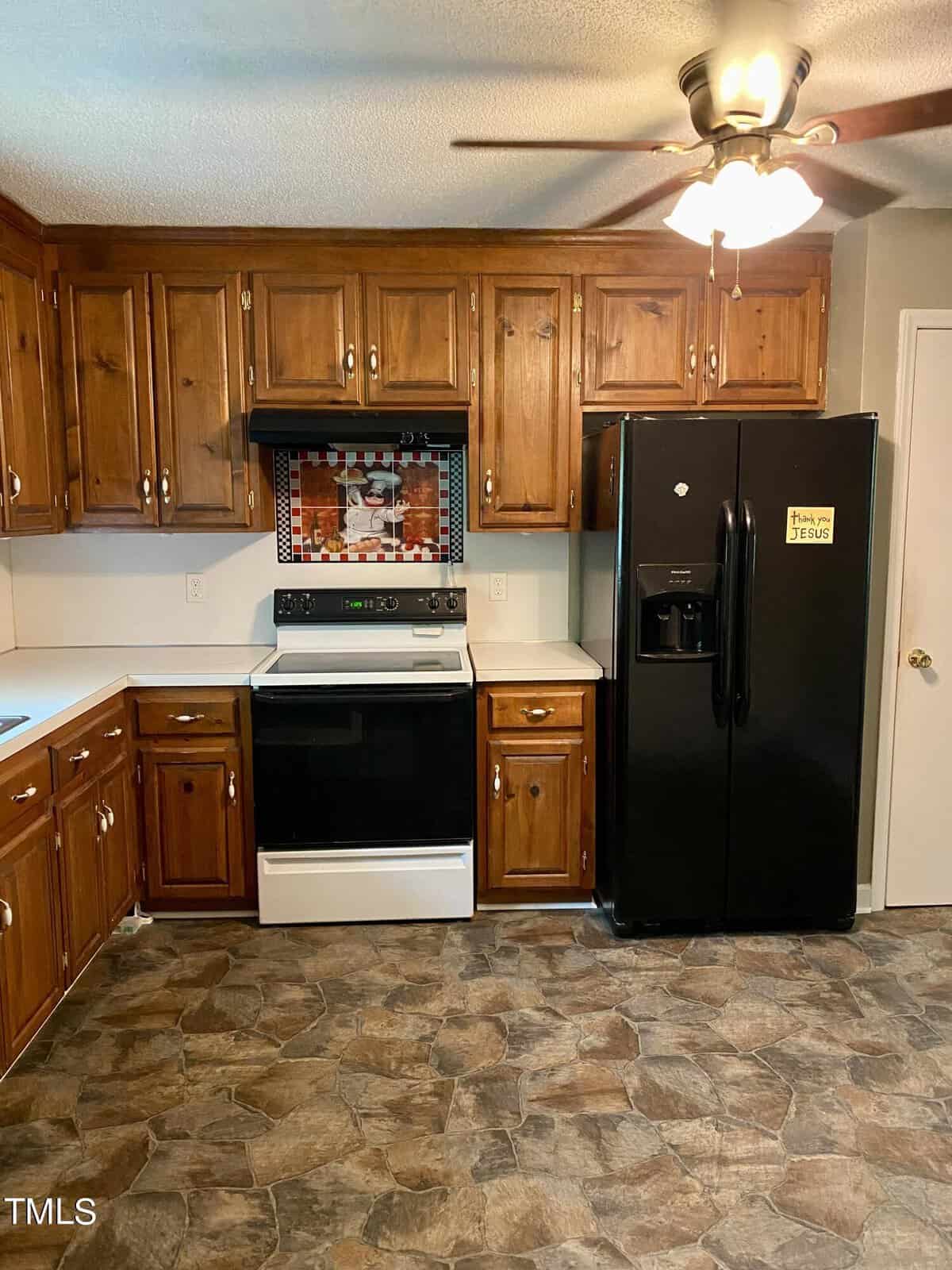 A kitchen with wooden cabinets, a white electric stove with a black cooktop, a black refrigerator, and a ceiling fan with a light. The backsplash behind the stove features a colorful tiled design. The floor is covered with stone-patterned vinyl.