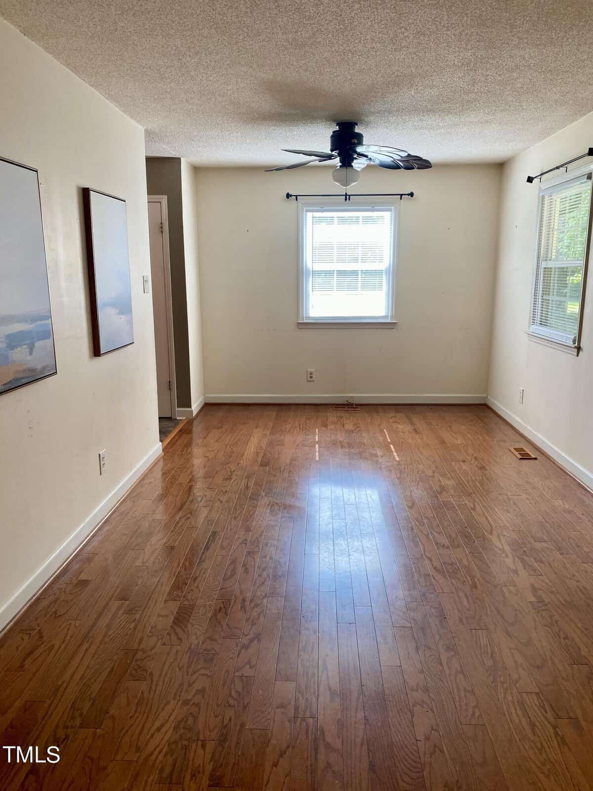 A vacant room with light-colored walls and hardwood floors. A ceiling fan is mounted in the middle of the ceiling. The room is illuminated by two windows: one on the far wall and one on the right wall. There are two picture frames on the left wall.