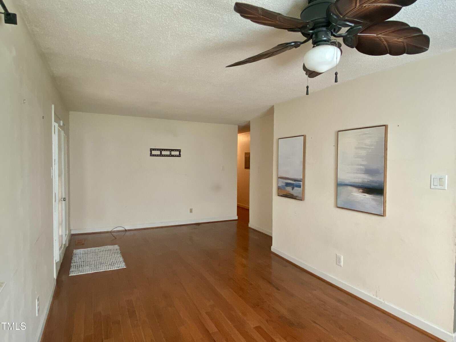 A sparsely furnished living room with wood flooring. There are two abstract paintings on the right wall and an empty TV wall mount on the back wall. A ceiling fan with large, leaf-like blades hangs from the ceiling, and light filters in from an unseen window.