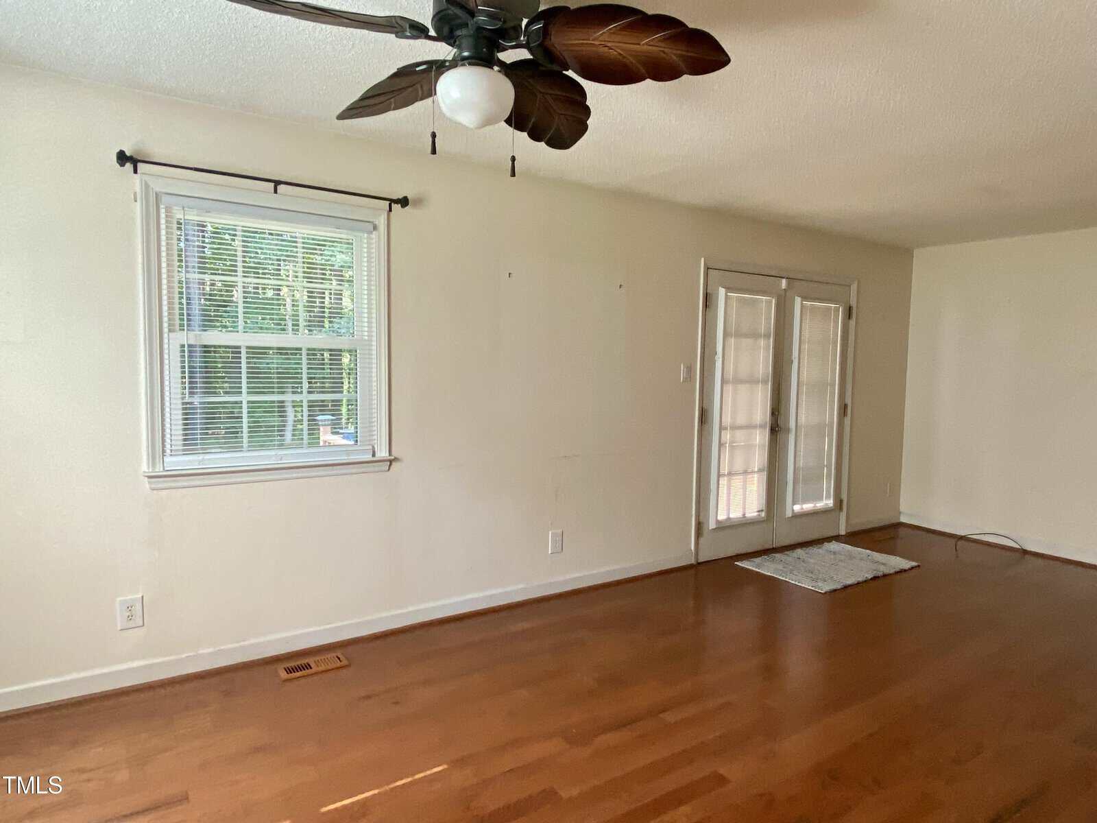 An empty room with hardwood flooring, a ceiling fan with leaf-shaped blades, a window with blinds on the left wall, and double French doors with glass panes on the back wall. There is a small rectangular rug near the French doors.